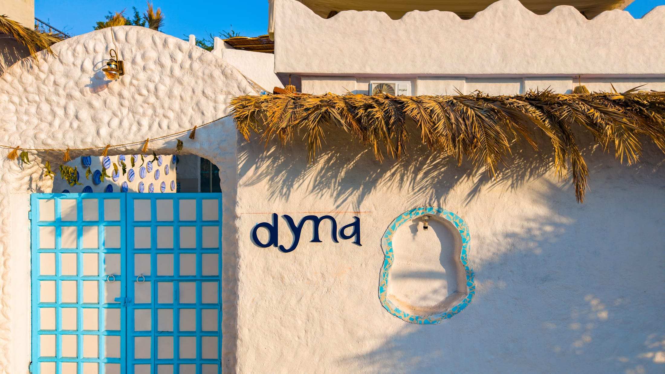Hand-painted Dyna sign on a textured white wall beside a blue lattice gate and thatched eave